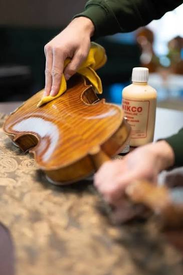 Manos limpiando con un paño amarillo la tapa barnizada de un violín, con un frasco de producto al fondo